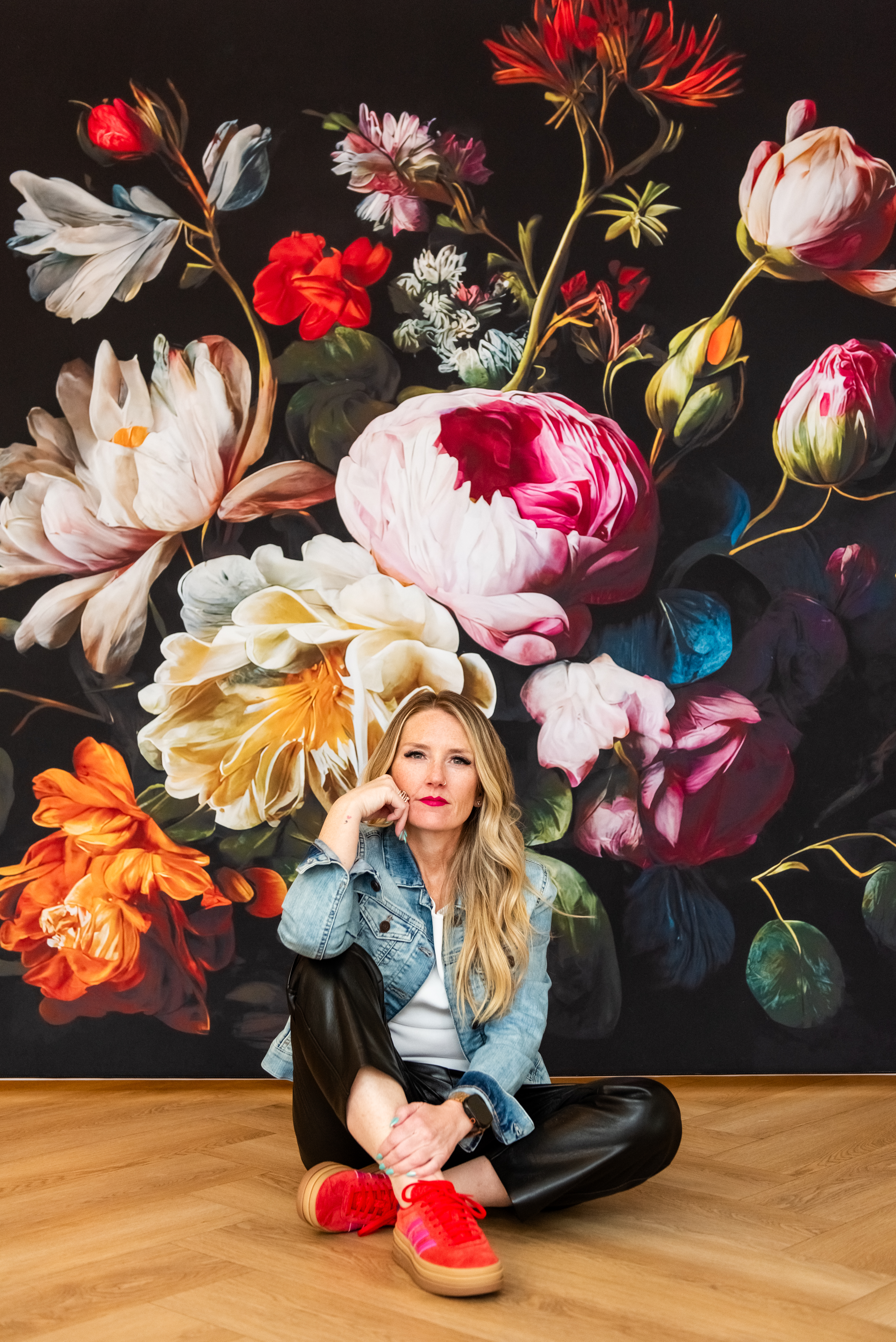 Alyson sitting in front of a vibrant floral mural, wearing a denim jacket and red shoes